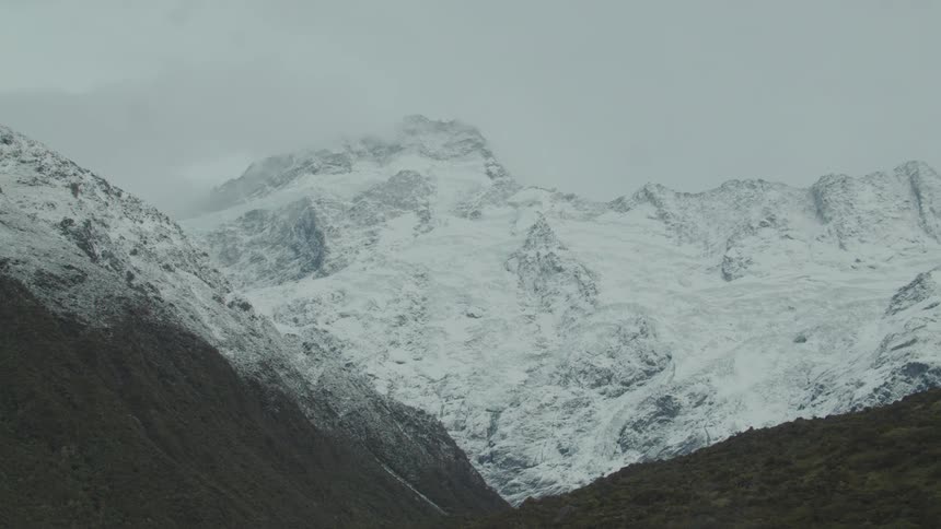 A massive snowcovered mountain range stands under a cloudy sky with dark forested slopes in the foreground