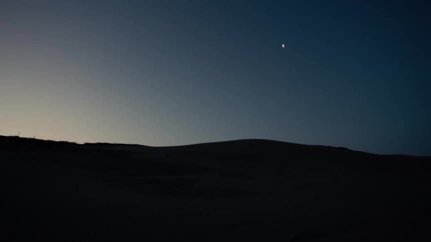 A thin crescent moon hangs in the darkening twilight sky above a silhouetted landscape of rolling hills or dunes