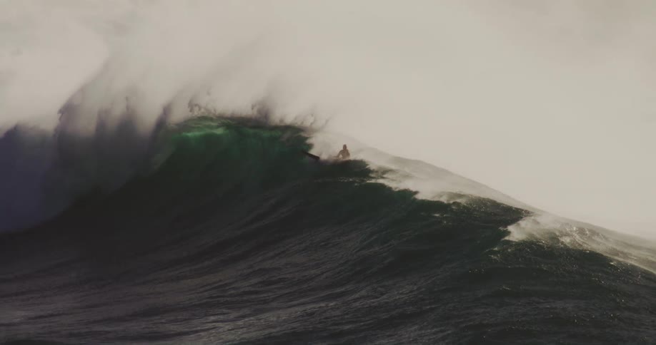 A surfer paddles atop a massive dark green wave as it begins to curl and break