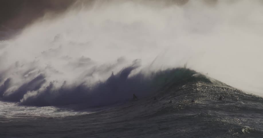 Several surfers are scattered across a large dark green wave as it crests with white spray rising from its peak
