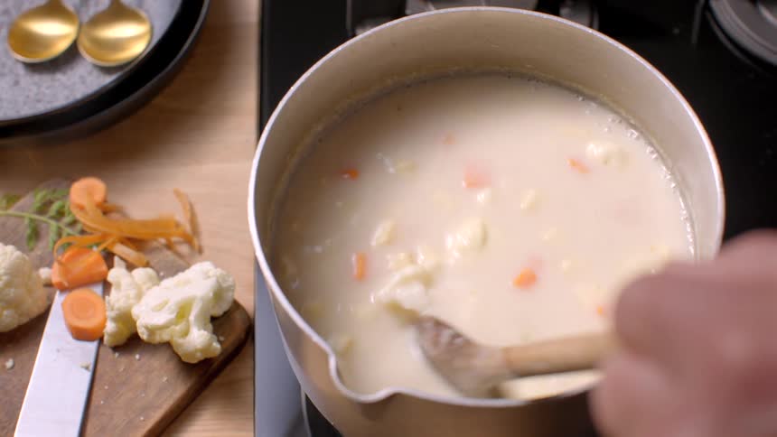 A person stirring soup on a stove with a wooden spoon while adding ingredients