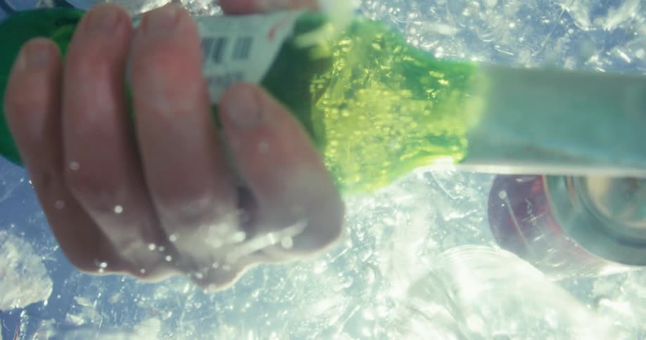 A person reached into an icefilled water cooler picking up a bottle of beer two cans floated nearby in the water at midday