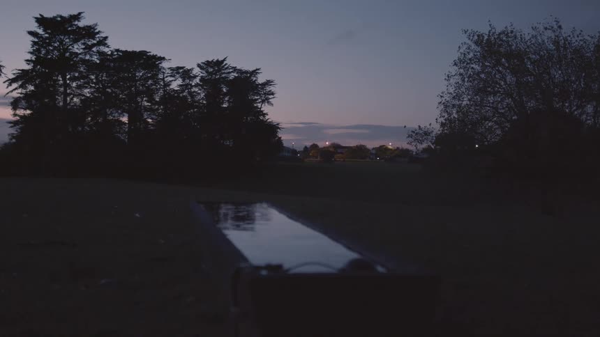A narrow reflective watering trough with silhouetted trees and distant lights against a dusky sky