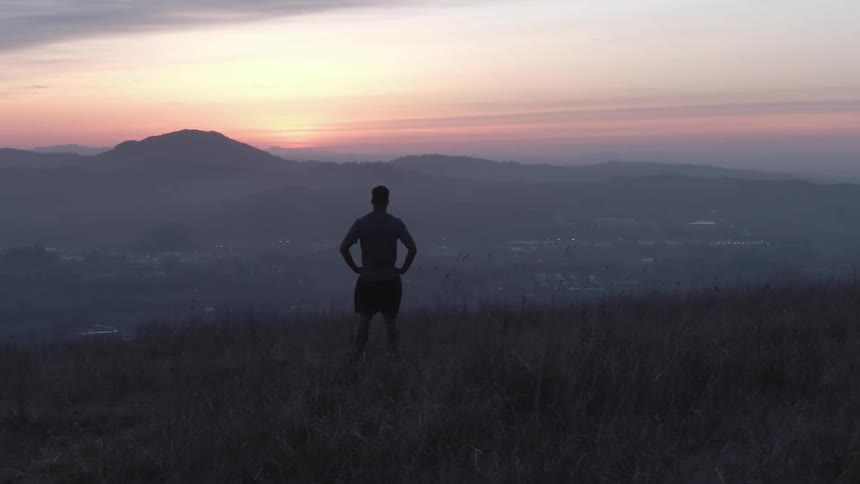 A silhouetted figure stands on a dark grassy hilltop facing a sweeping landscape of hazy mountains and a valley bathed in the soft colorful hues of a sunrise or sunset sky