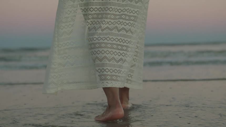 Close up of legs of a person in a white skirt walking in the water at the beach