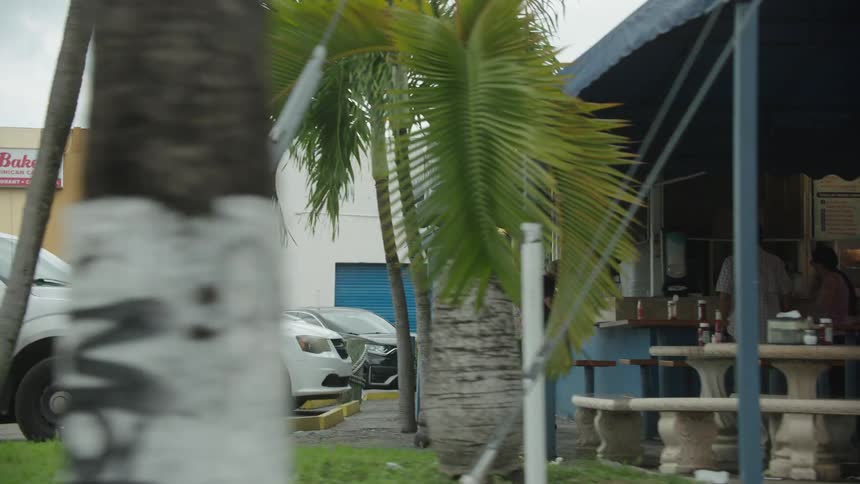 A group of people standing outside a restaurant placing orders with their cars parked along the side at midday