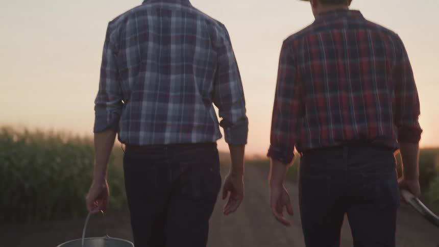 Two men in cowboy hats one carrying a bucket and the other a hoe walk away down a dirt path between fields as the sun sets behind them