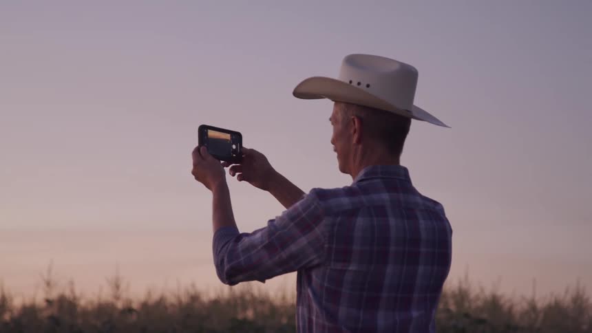 A man in a cowboy hat takes a photo with his phone in an open field at dusk