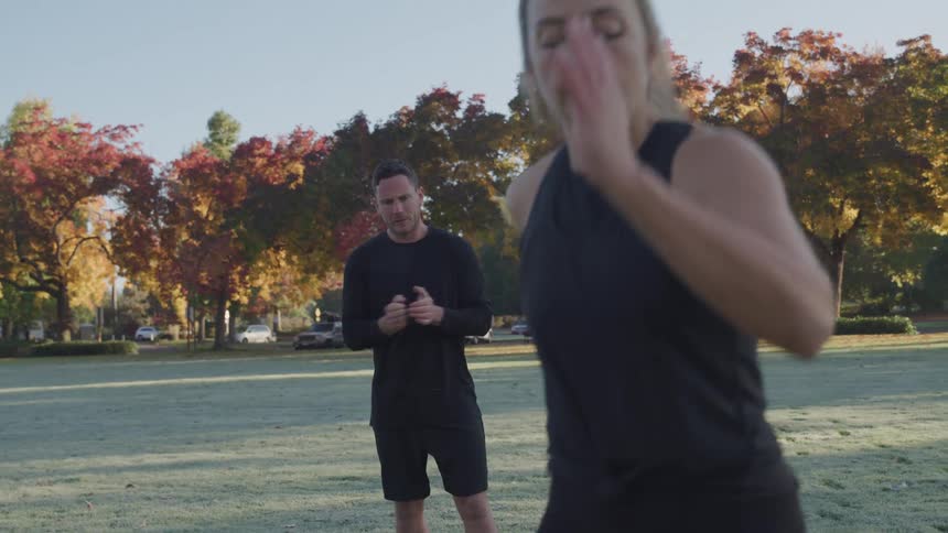A female runner in a black tank top is warming up with a male trainer looking on in a park filled with autumncolored trees under a clear sky
