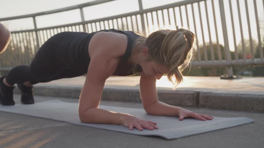 A blonde woman in black athletic wear is holding a plank position on a grey exercise mat on a concrete bridge with a persons hand on her back illuminated by the setting sun
