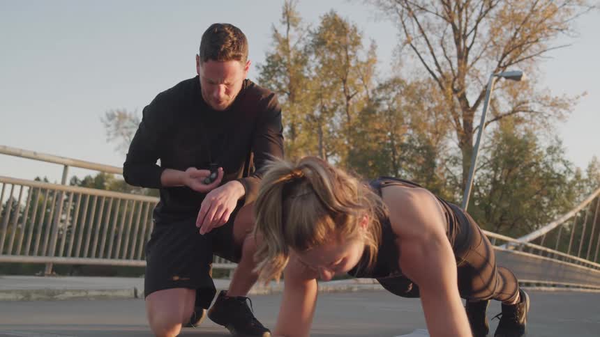 Trainer timing client as she performs a plank outdoors at a park