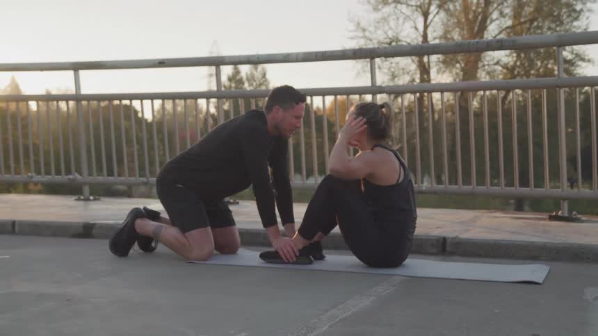 Woman does situps outdoors on a bridge during the day with the help of a trainee who holds her legs to the floor