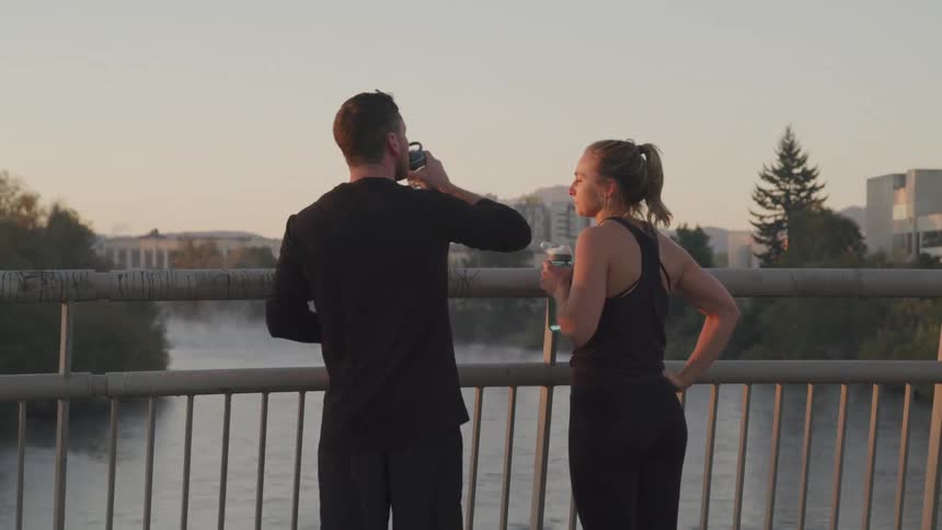 A man and woman in black athletic wear stand facing away from the viewer on a bridge overlooking a misty river and distant city skyline at sunset