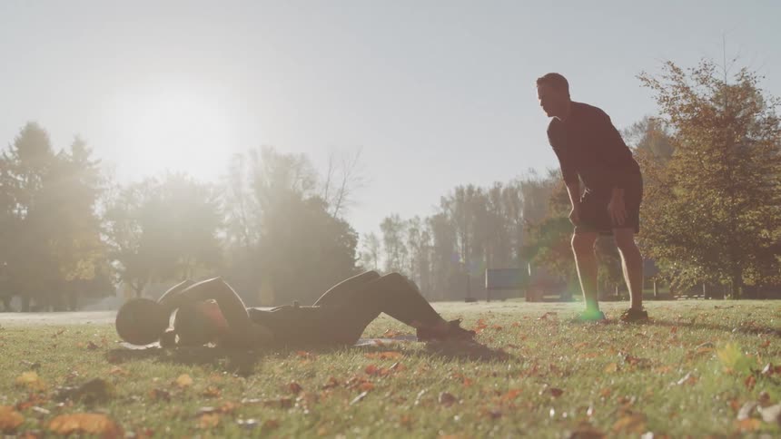 Man and woman throw a ball back and forth while the woman does sit ups on the grass