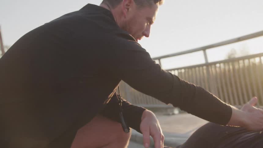 A male trainer in a dark longsleeved shirt holds a stopwatch glancing down at a woman doing a plank on a concrete bridge with sunlight in the background