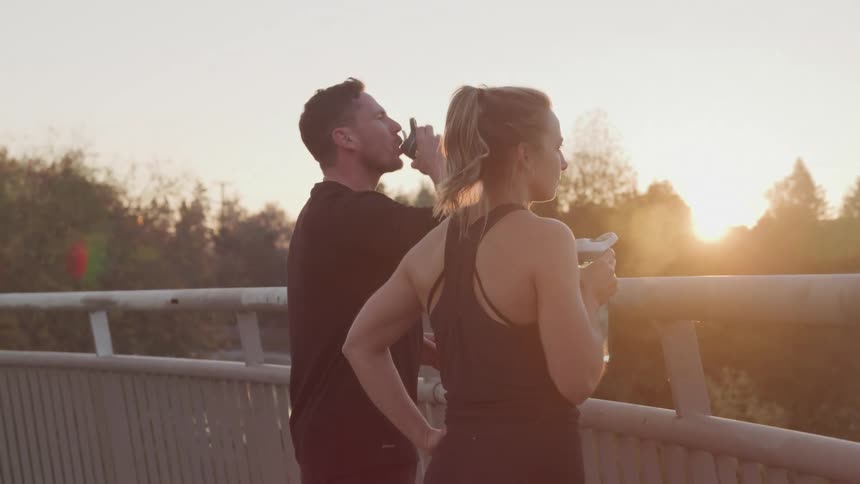 Young woman and her male trainer drink water from water bottles while standing on a bridge during the day