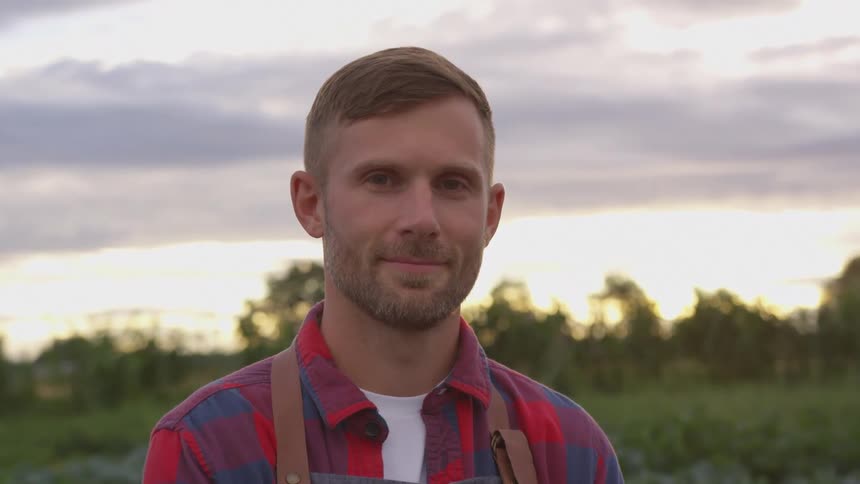 A young man with short hair and a beard wearing a red and blue plaid shirt stands against a blurred background of a field and a cloudy sky