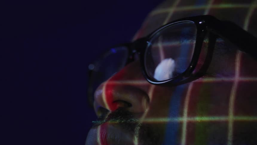 A man wearing black glasses has a red and blue grid pattern projected onto his face in a dark setting