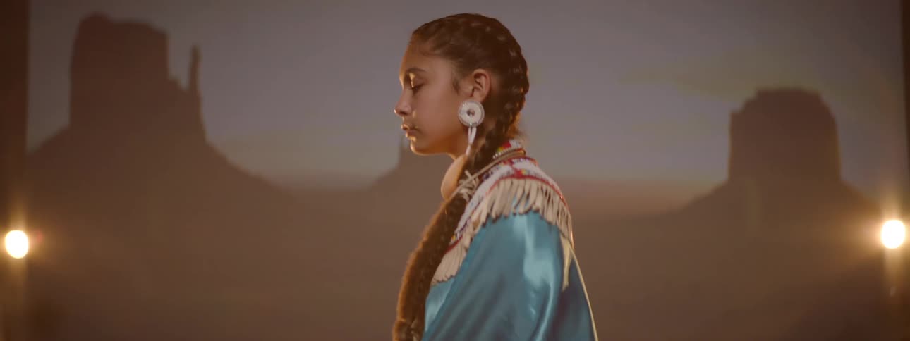 A teenage girl in traditional indigenous attire including a fringed blue and white top and decorative earrings stands with their eyes closed against a background featuring silhouetted rock formations and stage lights
