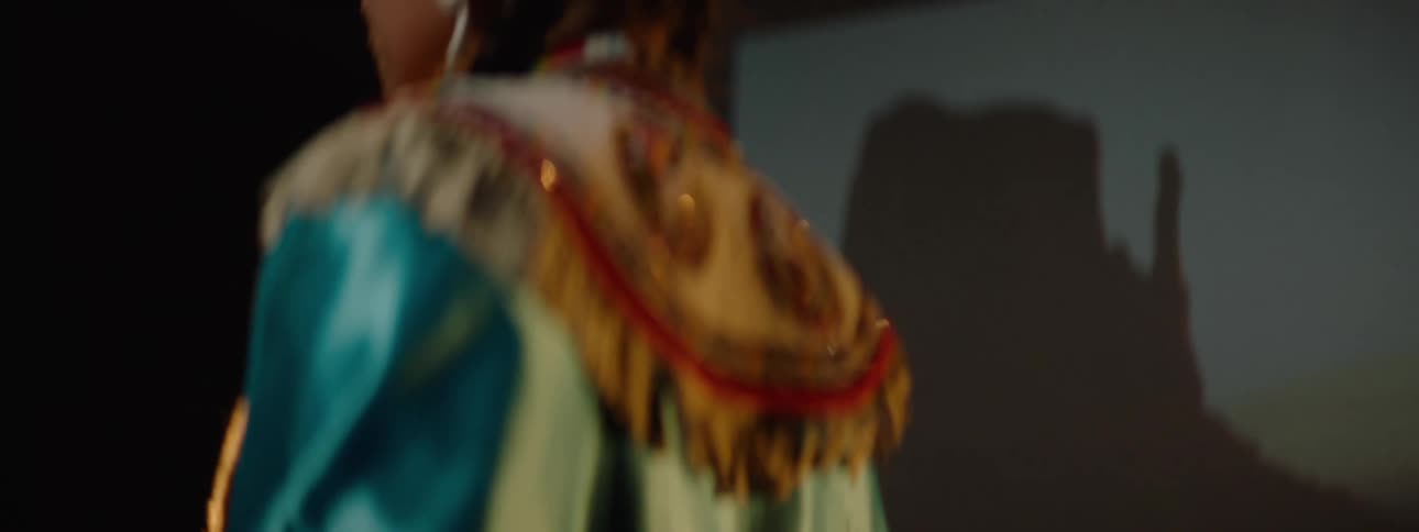 A native american teenager wearing a satin traditional outfit stands on a stage