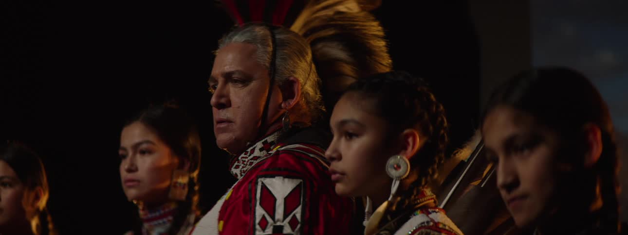 A group of people stands on a stage including a man and four young women all dressed in traditional american indian attire