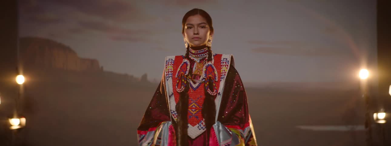 A young woman in native regalia stands proudly under stage lights