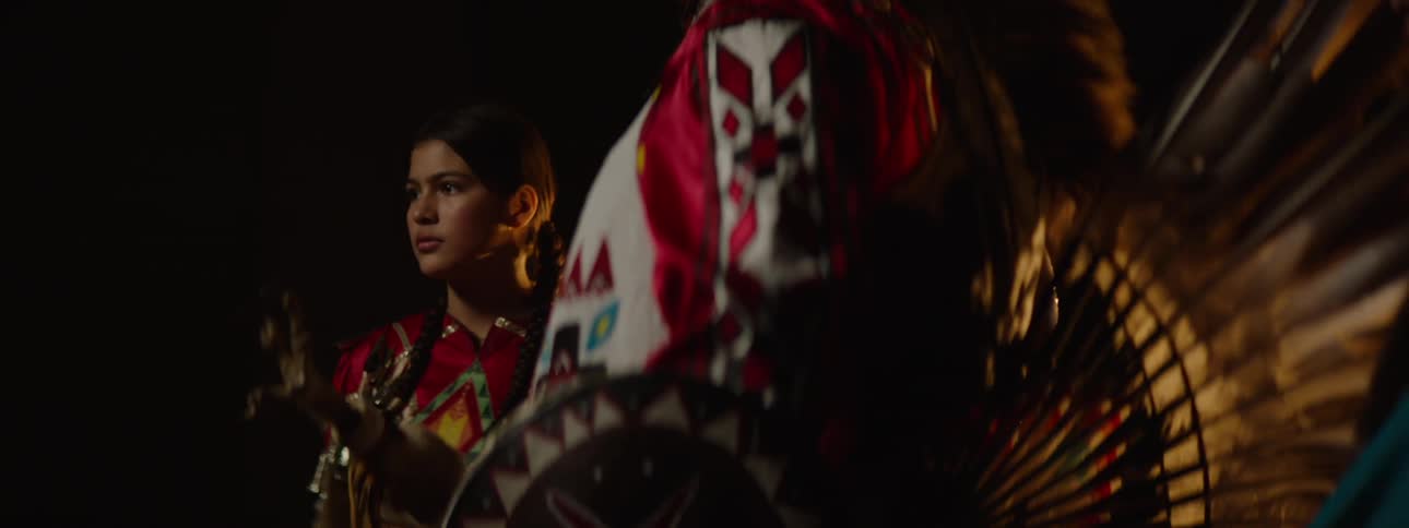 A group of native americans dressed in colorful traditional attire stands together on a dimly lit stage