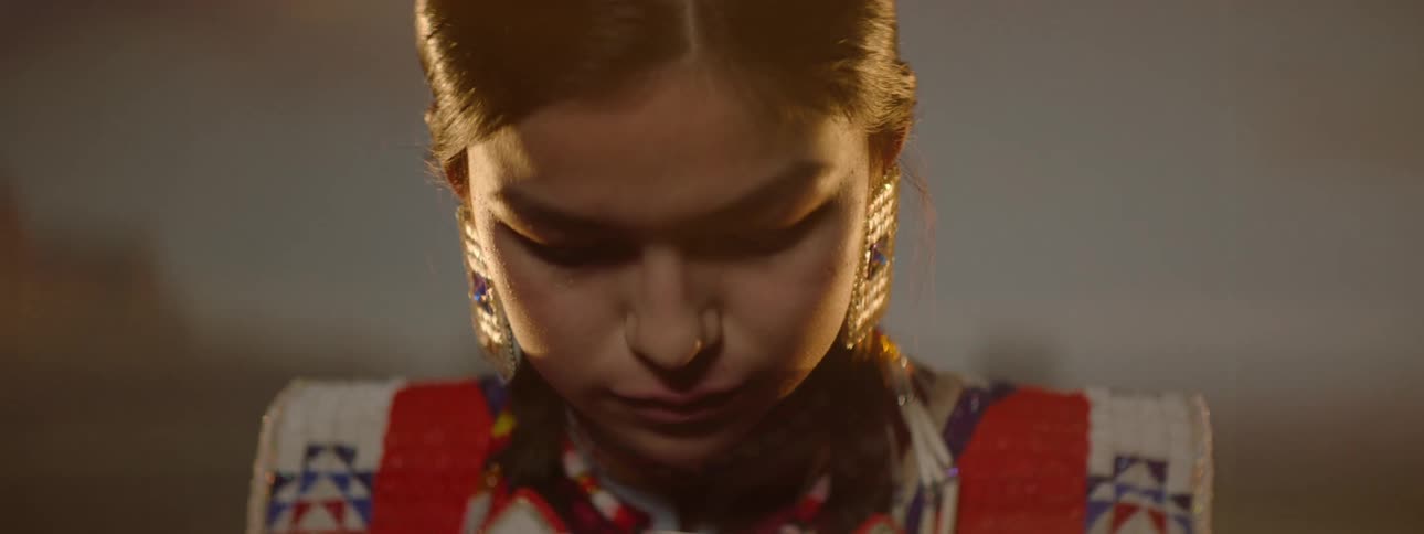 A native american teenage girl wearing traditional attire adorned with jewelry and makeup stands confidently on a stage