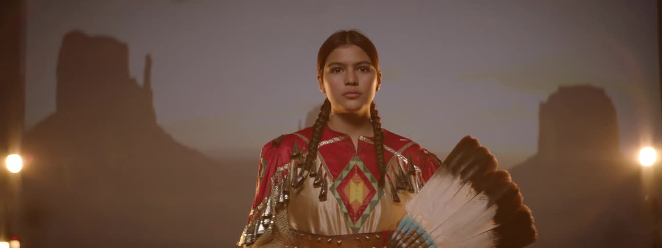 A native american teenage girl stands on a stage wearing traditional attire holding a feather fan partially over her face