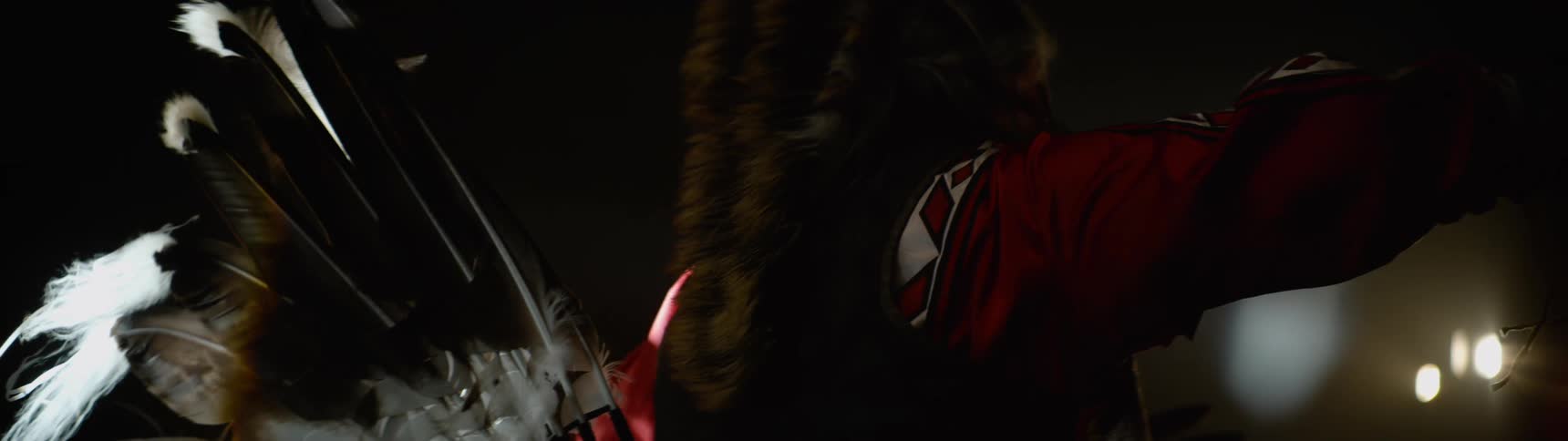 A figure in elaborate indigenous regalia featuring prominent light colored feathers fur and a red patterned garment shifts or prepares in a dimly lit space their attire catching highlights from a focused light source