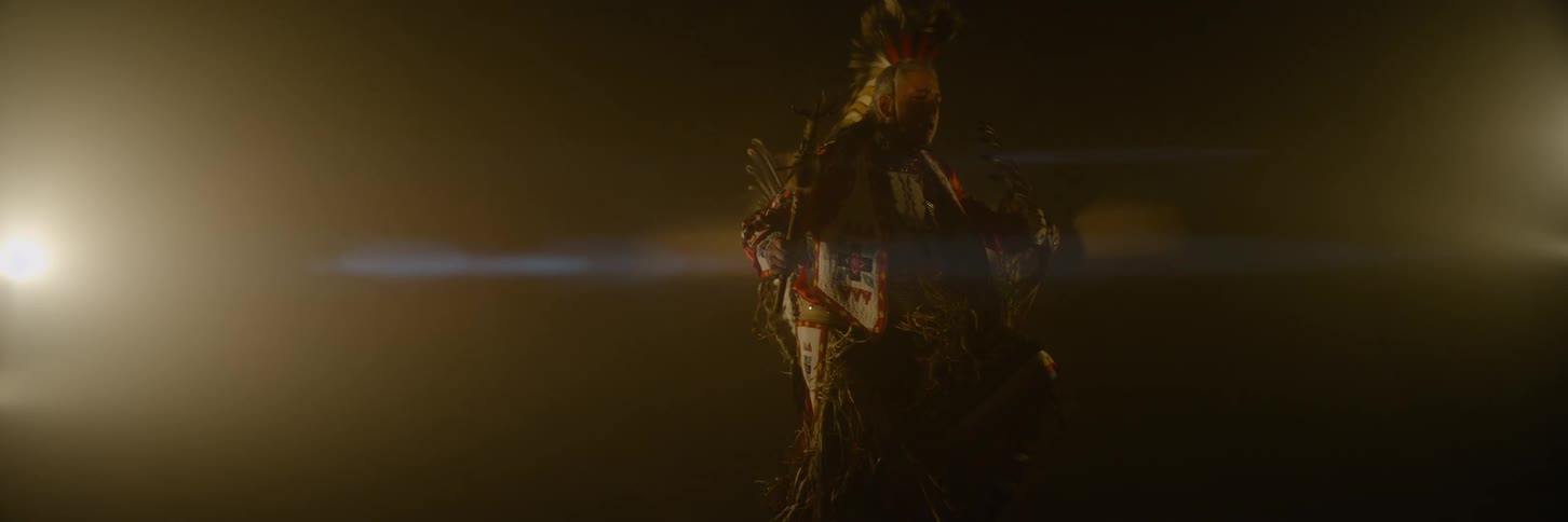 Older native american male dances while wearing ceremonial attire in an area lit by a bright light