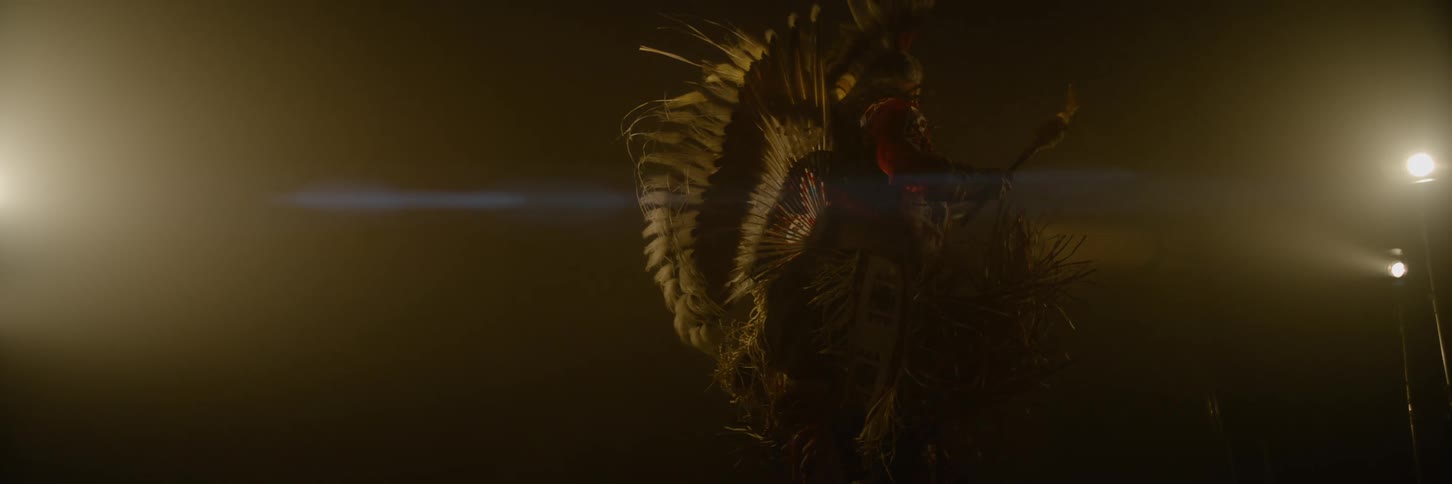 A person in elaborate traditional indigenous native american regalia adorned with feathers and fringe performs a dance or ritual in a dark atmospheric setting with warm low lighting and a visible lens flare
