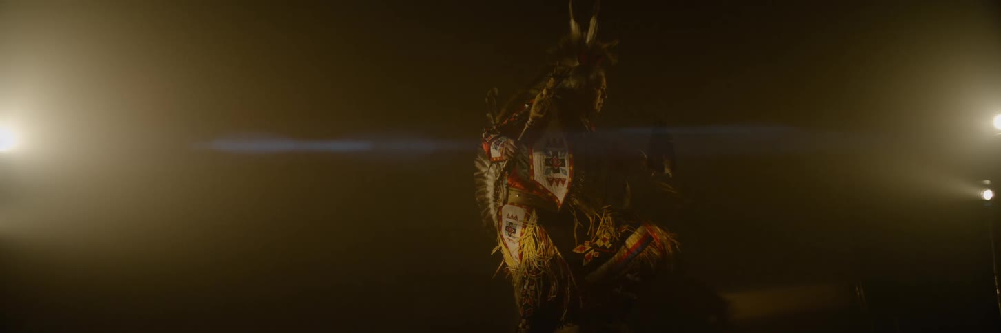 A native american man dressed in traditional feathered attire dances under warm dim lighting