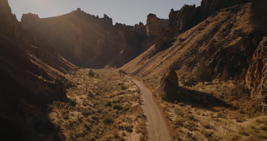Aerial view gliding over a winding dirt road through a dry desert canyon surrounded by rugged cliffs and rocky hills under warm sunlight