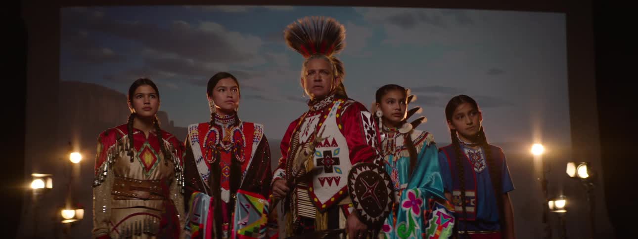 A group of native american indians including a man and several teenage girls all dressed in traditional attire standing on a stage