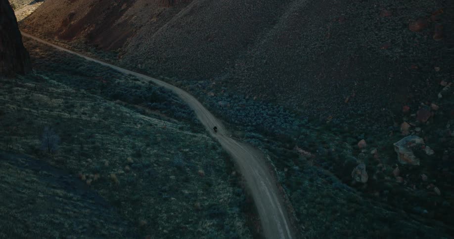 An aerial view shows a dirt road winding through a deep shadowed canyon with steep rocky slopes and sparse vegetation with a motorcycle traveling on it under a sunlit sky