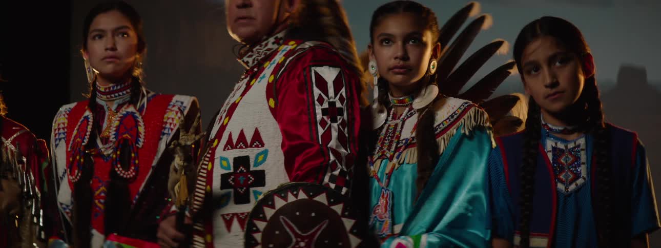 Native americans in traditional regalia stand solemnly under dramatic lighting