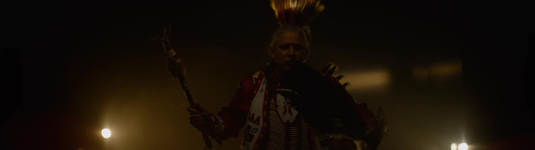 In a dimly lit and hazy setting a man in elaborate feathered indigenous regalia holds a staff and performs a ceremonial dance with dramatic light catching his face and attire