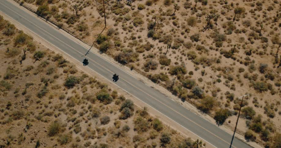 Aerial view of two motorcycles traveling along a paved road surrounded by a flat area with sparse vegetation