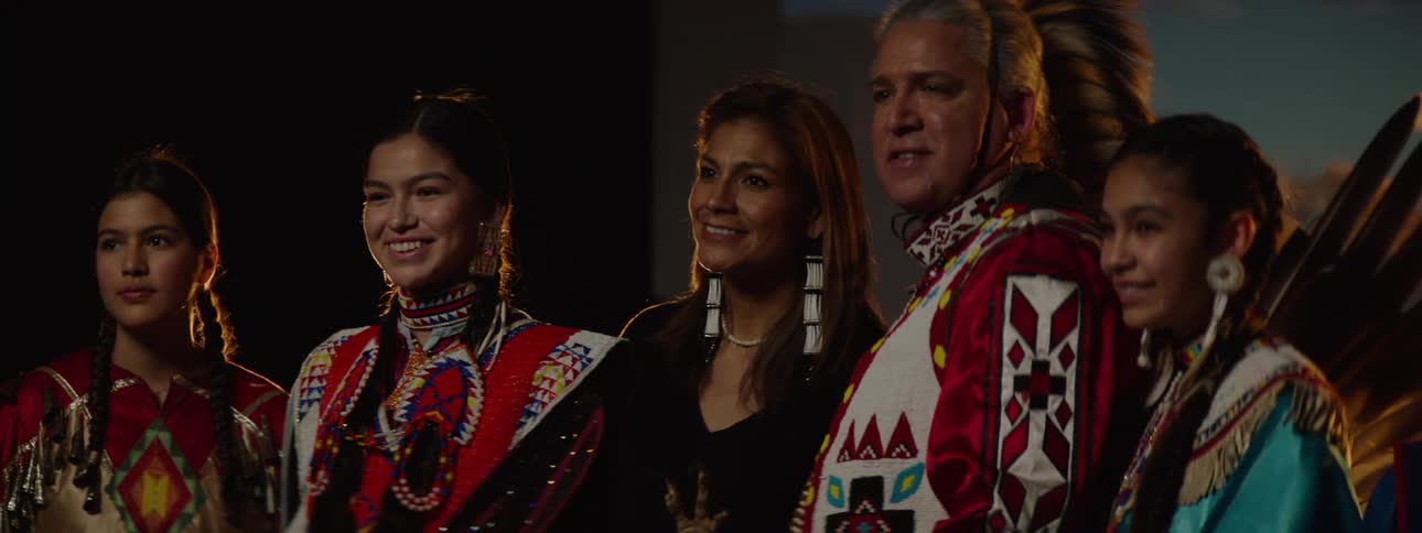 A group of native american individuals are standing sidebyside in traditional regalia against a dark background