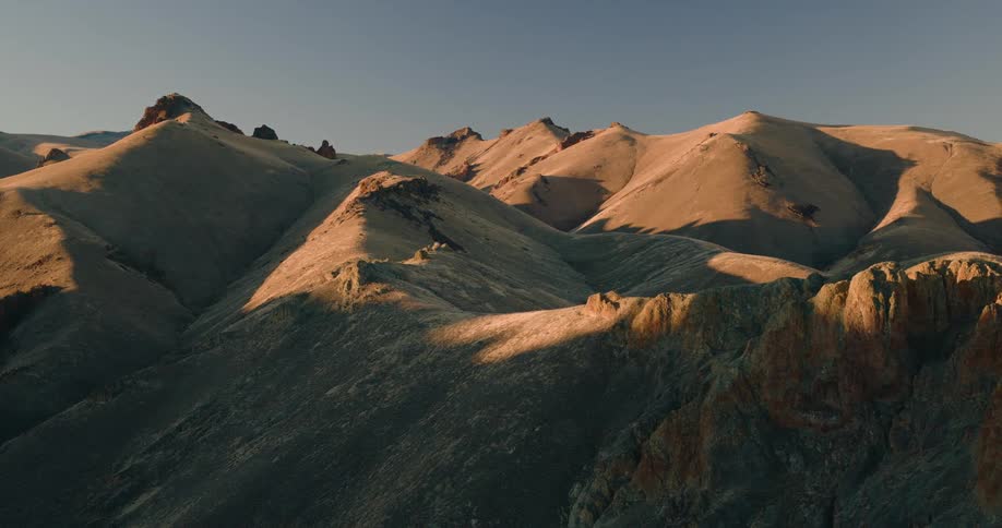 Golden sunlight grazes the rugged peaks of barren mountains in an arid canyonlike setting