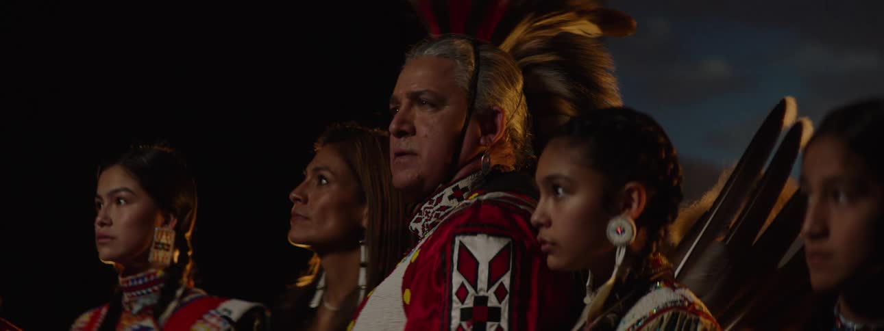 A group of people wearing traditional native american clothing and accessories including feathered headdresses and beaded earrings are gathered together