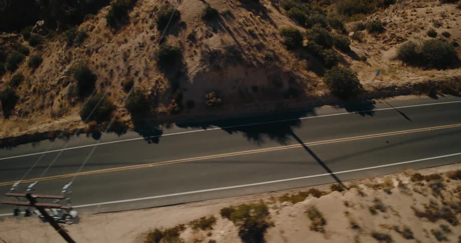 An aerial view tracks a twolane road as it winds through a dry rocky desert landscape bordered by scattered brush and rugged hills