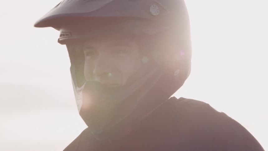 A person wearing a motorcycle helmet stands backlit by the sun partially silhouetted