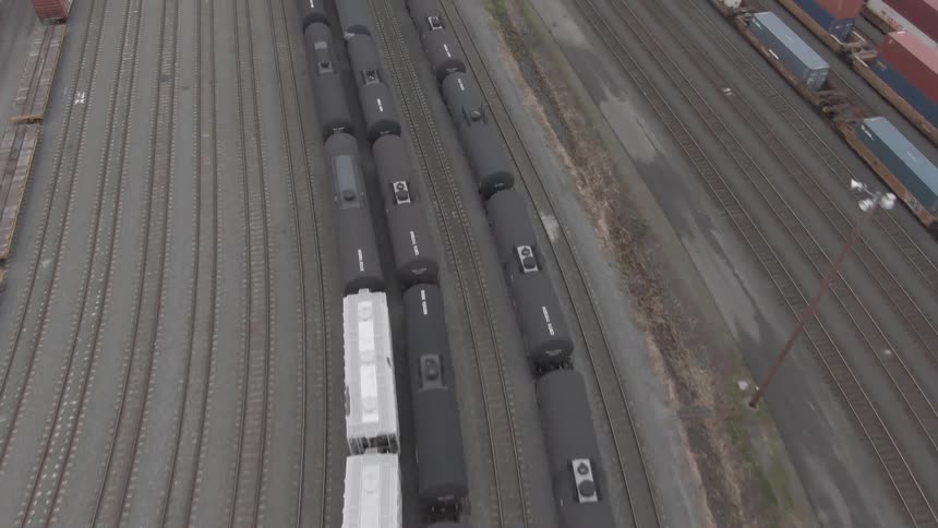 An aerial view flies over a large rail yard filled with many parallel tracks and long lines of tanker and freight cars