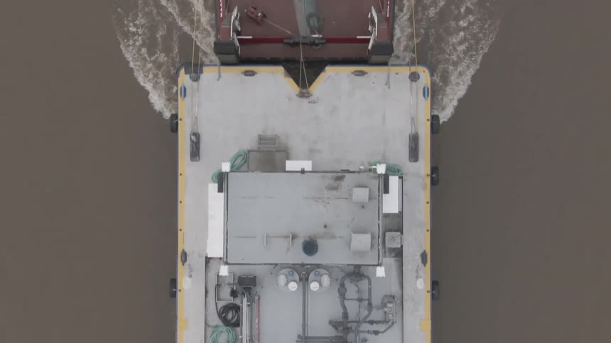 An aerial view captures a tugboat towing a large vessel along a winding river leaving a wake behind as it cuts steadily through the water