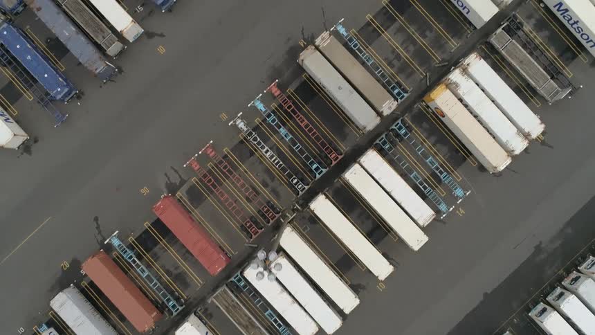 An overhead shot displays a large paved lot filled with various rectangular shipping containers and empty transport dollies