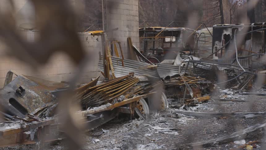 Ashy remnants of a property that has been destroyed by a fire are viewed through a chain link fence