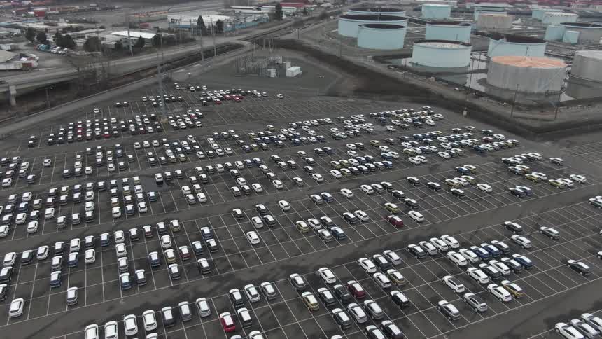 A large parking lot filled with parked cars next to a factory under the bright midday sun