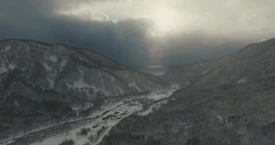 An aerial view captures a wide snowcovered valley with a winding dark river surrounded by mountains and a cloudy sky with sunlight breaking through